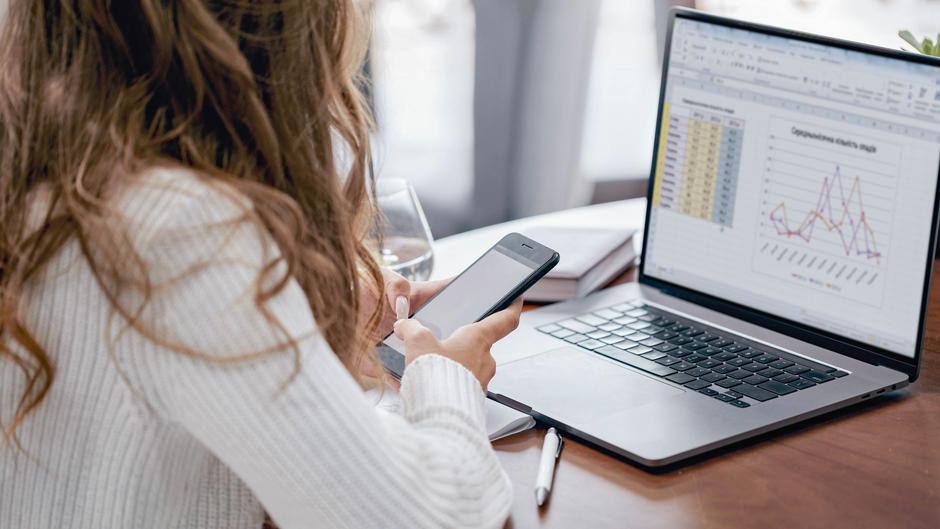 Eine Person mit lockigem Haar sitzt an einem Tisch und nutzt ein Smartphone, während ein Laptop mit Daten und Diagrammen geöffnet ist.