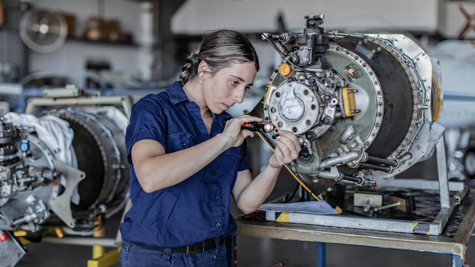 Eine Mechanikerin arbeitet konzentriert an einem Flugzeugmotor und beleuchtet ihn mit einer Taschenlampe.