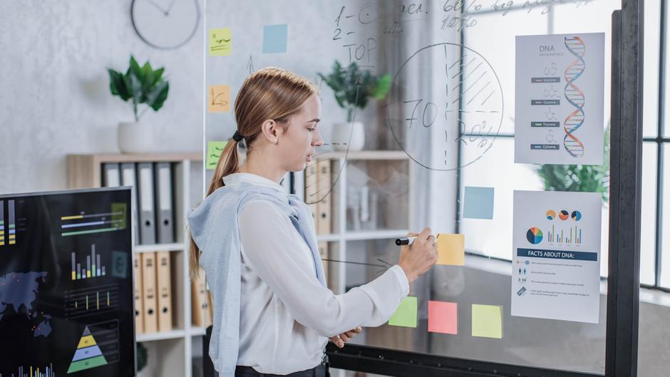 Eine Person schreibt mit einem Marker auf eine transparente Tafel, umgeben von bunten Haftnotizen und Informationsblättern.
