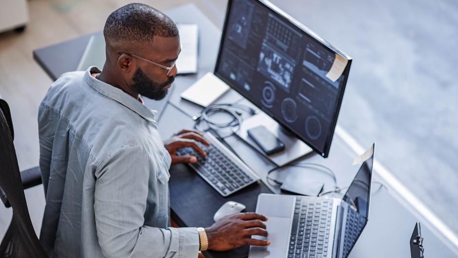 Mann arbeitet an mehreren Computern mit Monitoren und Tastaturen in einem modernen Büro.