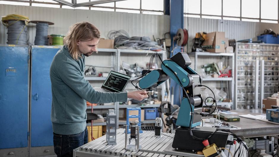 Ein Mann arbeitet an einem Roboterarm und hält ein Bauteil, während ein Laptop auf dem Tisch steht.