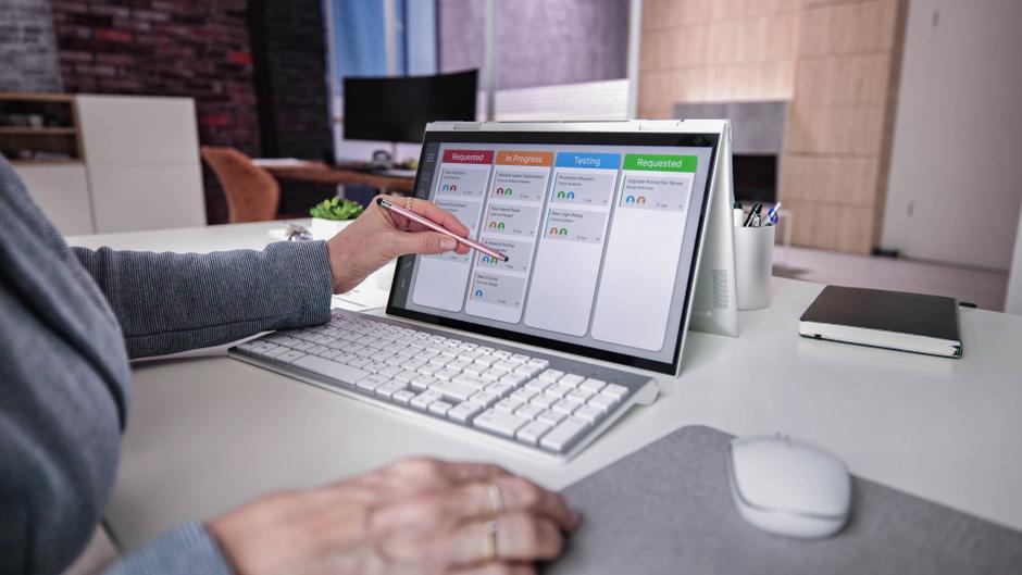 Eine Hand mit einem Stift zeigt auf ein Tablet, das eine Projektmanagement-Übersicht mit verschiedenen Statusanzeigen zeigt.