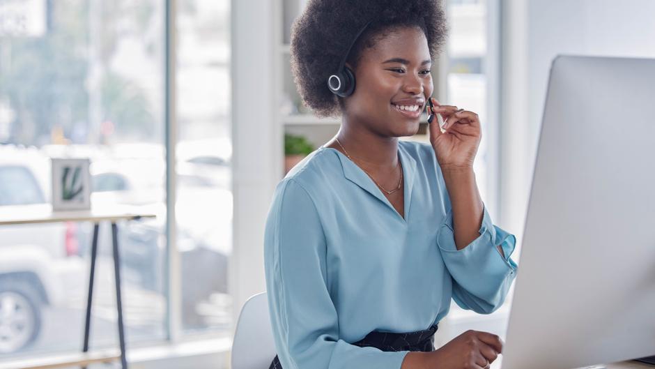 Lächelnde Frau mit Headset sitzt vor einem Computer und spricht in ein Mikrofon.