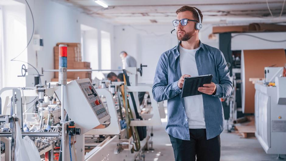 Mann mit Kopfhörern und Tablet in einer Produktionshalle, Maschinen und Arbeiter im Hintergrund.