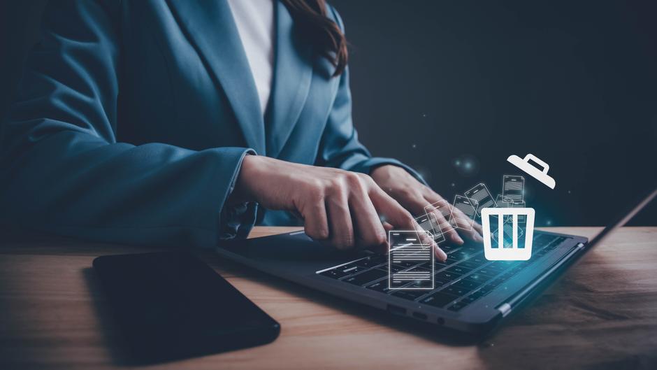 Eine Person in einem blauen Blazer tippt auf einer Laptop-Tastatur, umgeben von digitalen Icons von Dokumenten und einem Papierkorb.