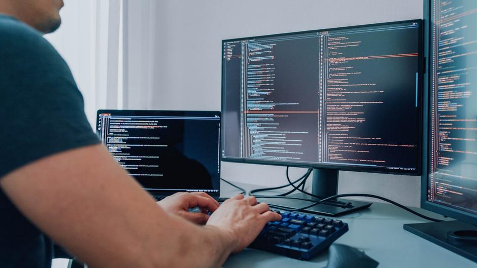 Hände tippen auf einer Tastatur, während zwei Computerbildschirme mit Codezeilen angezeigt werden.