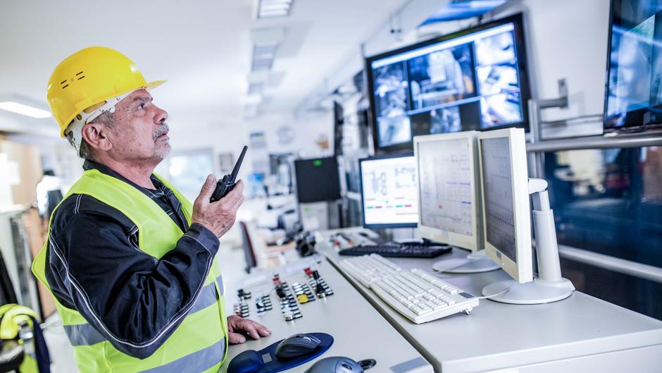 Ein Mann mit gelbem Helm und Warnweste spricht mit einem Funkgerät vor mehreren Monitoren in einem Kontrollraum.