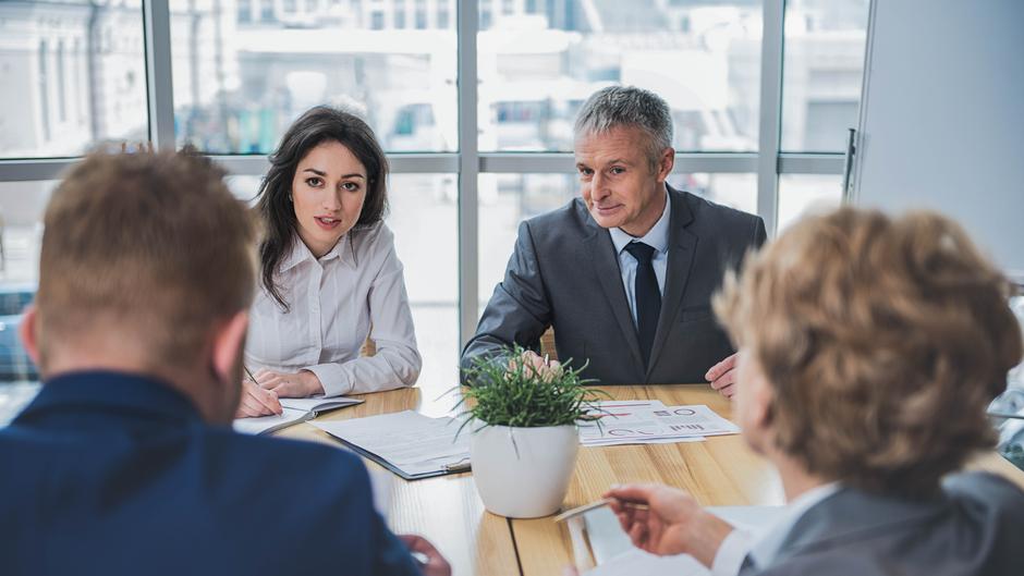 Vier Personen in formeller Kleidung sitzen an einem Tisch, diskutieren und schauen auf Dokumente. Ein Pflanzenarrangement steht in der Mitte.