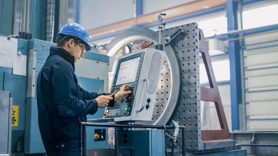 Ein Arbeiter in einem blauen Helm bedient eine CNC-Maschine in einer industriellen Umgebung.
