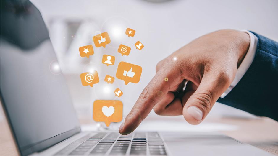 Eine Hand tippt auf eine Laptop-Tastatur, während orangefarbene Symbole für soziale Medien darüber schweben.