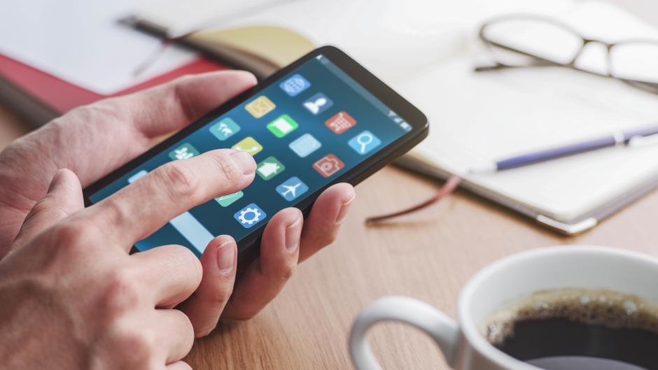 Eine Hand tippt auf ein Smartphone mit verschiedenen App-Symbolen, im Hintergrund eine Tasse Kaffee und ein Notizbuch.