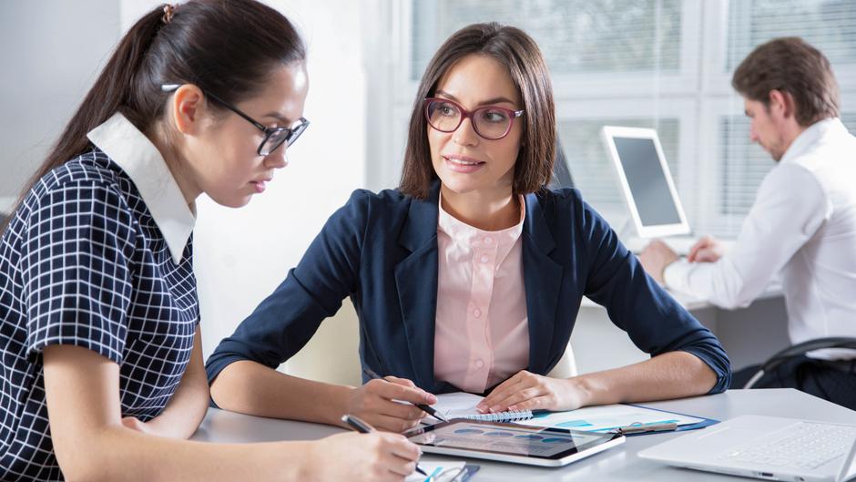 Zwei Frauen in einem modernen Büro besprechen Unterlagen, während ein Mann am Computer arbeitet.