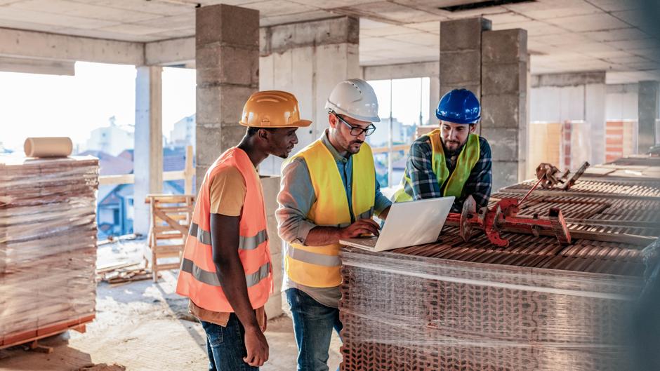 Drei Bauarbeiter in Sicherheitswesten und Helmen schauen auf einen Laptop, während sie auf einer Baustelle stehen.