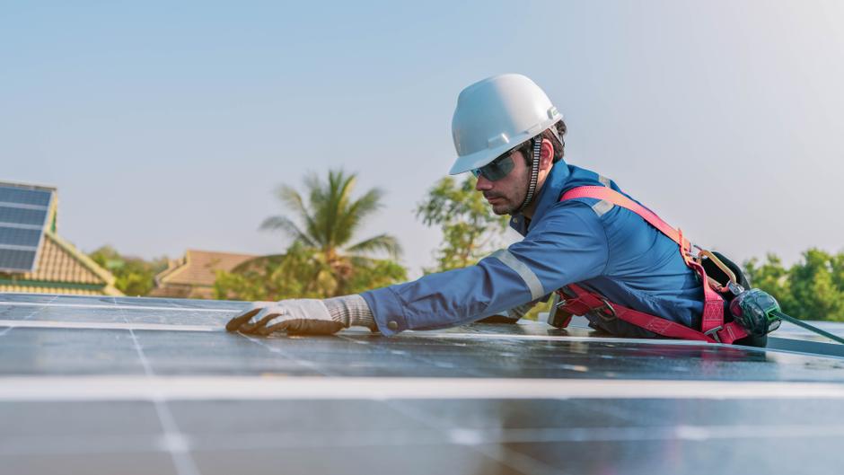Arbeiter in Schutzkleidung installiert Solarpanels auf einem Dach, umgeben von Bäumen und weiteren Gebäuden.