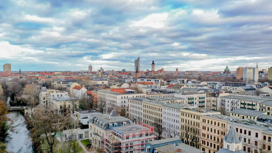 Stadtansicht mit historischen und modernen Gebäuden, Bäumen und einem Fluss unter bewölktem Himmel.