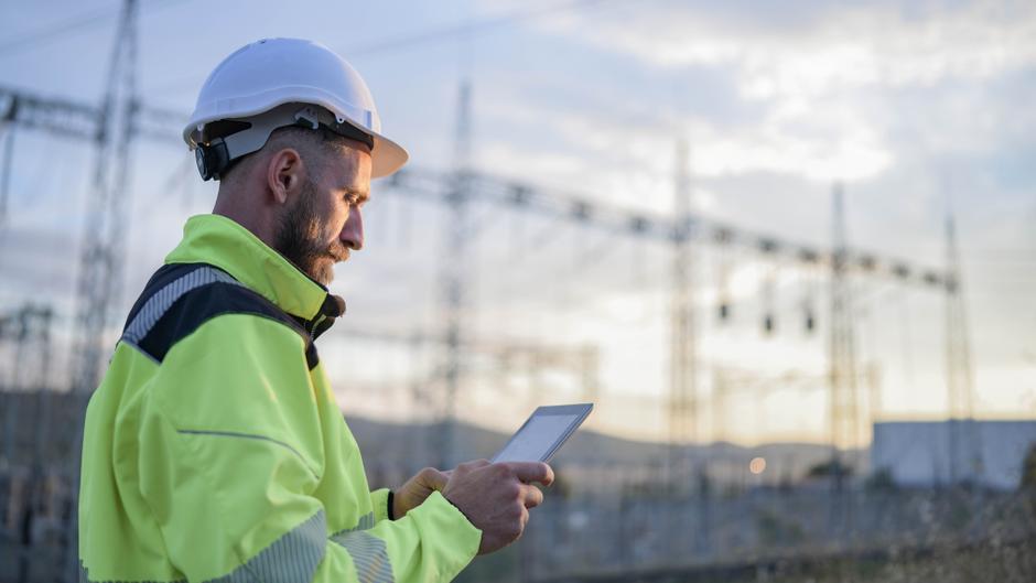 Ein Arbeiter in leuchtend gelber Sicherheitskleidung und Helm betrachtet ein Tablet vor einem elektrischen Umspannwerk.