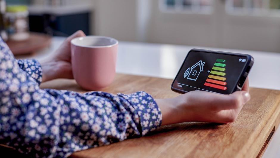 Eine Hand hält ein Smartphone mit einem Energieeffizienz-Display, während die andere Hand eine Tasse hält.