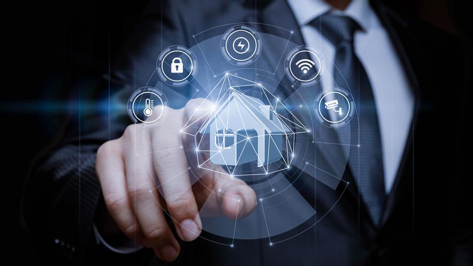 Hand eines Mannes in Anzug, die auf ein holografisches Haus mit verschiedenen Smart-Home-Symbolen zeigt.