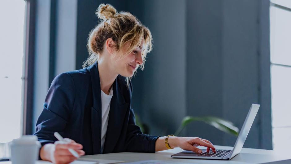 Frau mit lockigem Haar arbeitet lächelnd an einem Laptop in einem modernen Büro.