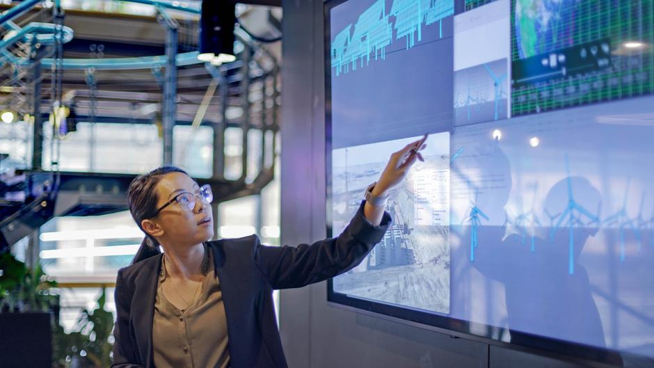 Eine Person mit Brille zeigt auf einen großen Bildschirm mit Grafiken und Diagrammen.