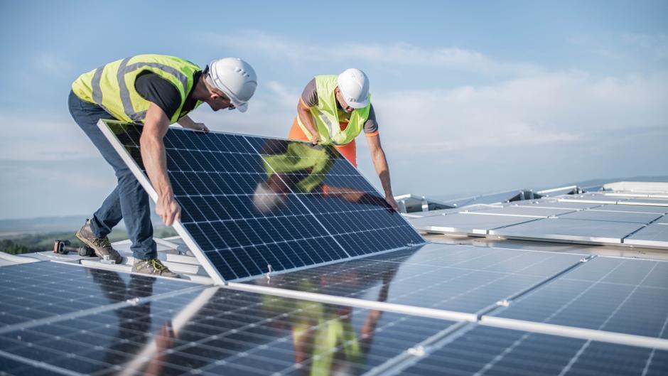 Zwei Arbeiter montieren Solarpaneele auf einem Dach, beide tragen Helme und Sicherheitswesten.