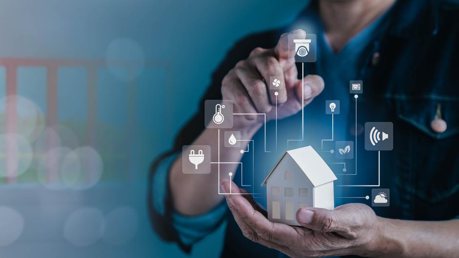Eine Person hält ein kleines Modellhaus und zeigt auf digitale Symbole für Smart-Home-Funktionen.