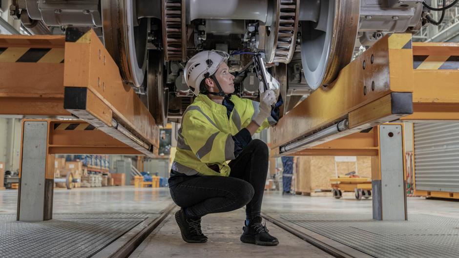 Eine Person in Sicherheitskleidung und Helm arbeitet mit einem Werkzeug unter einem großen Maschinenbauteil.