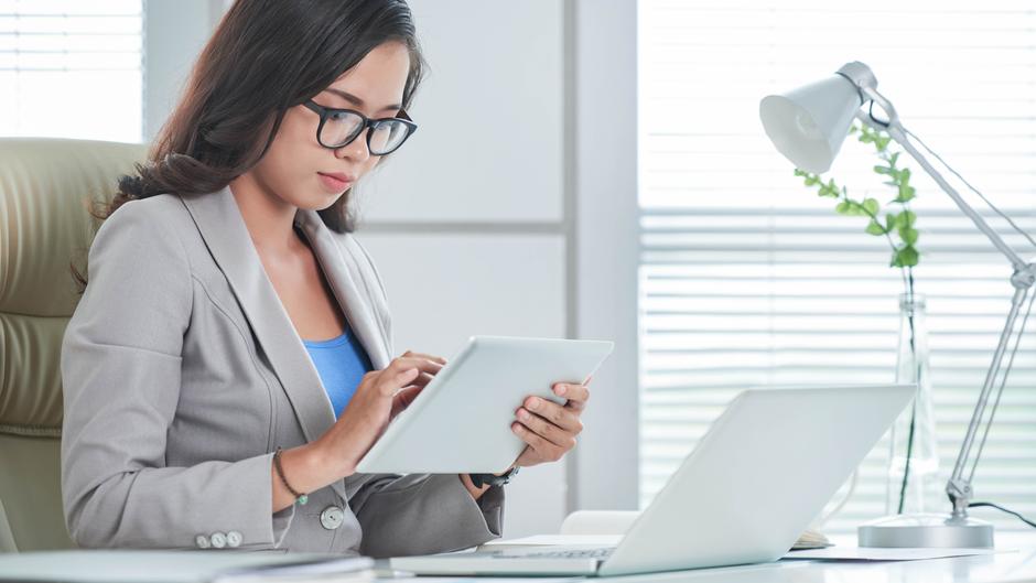Eine Frau in einem grauen Blazer sitzt an einem Schreibtisch und nutzt ein Tablet.