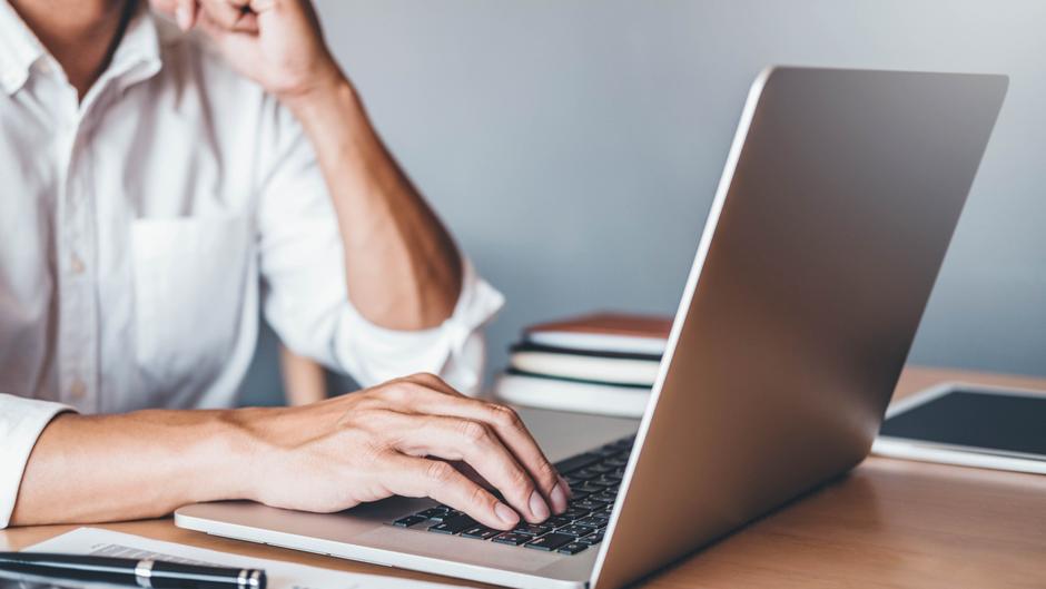 Eine Person tippt mit einer Hand auf einem Laptop, während die andere Hand das Kinn stützt.