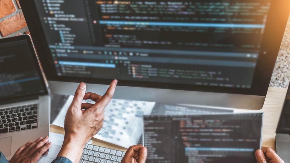 Hände zeigen auf einen Bildschirm mit Programmiercode, daneben ein Laptop und Notizen.