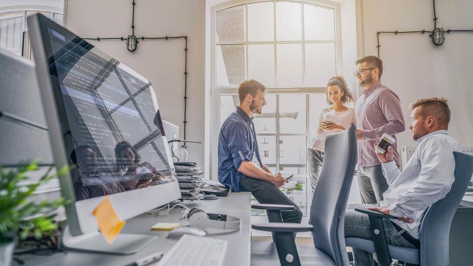 Vier Personen stehen und sitzen in einem modernen Büro, während sie miteinander sprechen und Smartphones nutzen. Ein Computerbildschirm zeigt Code.