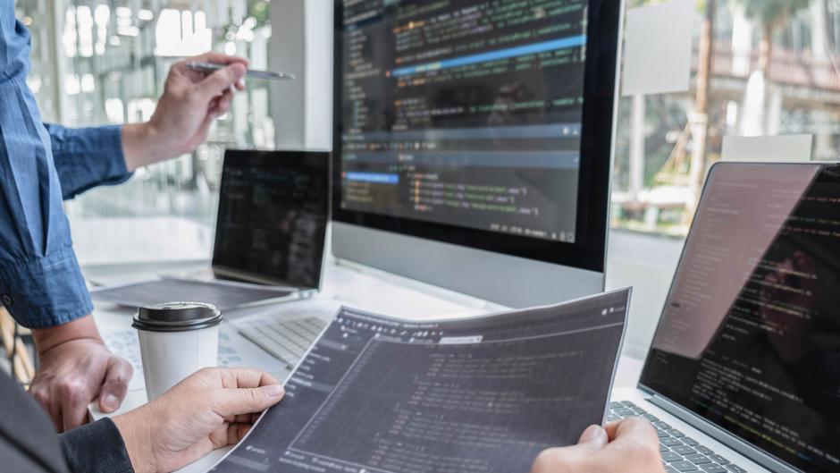 Zwei Personen diskutieren über Programmiercode, während sie auf mehrere Laptops und einen großen Monitor schauen.