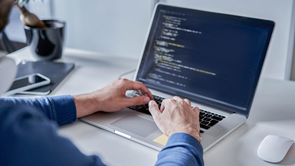 Hände tippen auf einer Laptop-Tastatur, auf dem Bildschirm sind Programmierzeilen zu sehen.