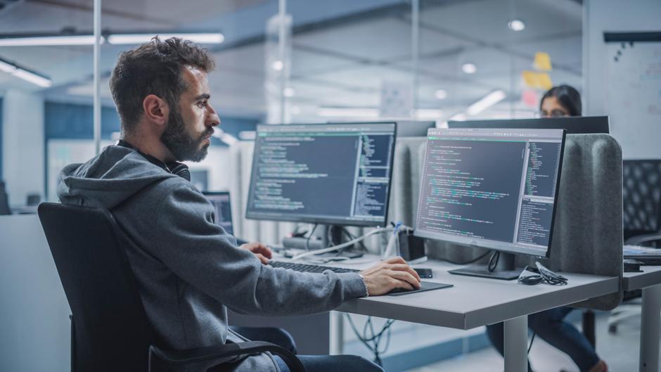 Ein Mann sitzt an einem Schreibtisch mit zwei Monitoren, auf denen Programmiercode angezeigt wird.