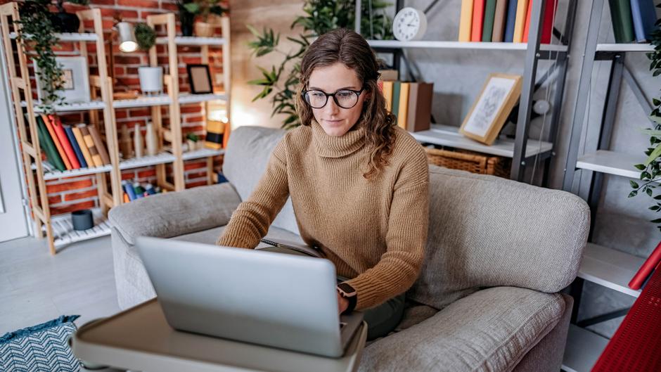 Frau mit Brille sitzt auf einem Sofa und arbeitet am Laptop in einem gemütlichen Raum mit Regalen im Hintergrund.