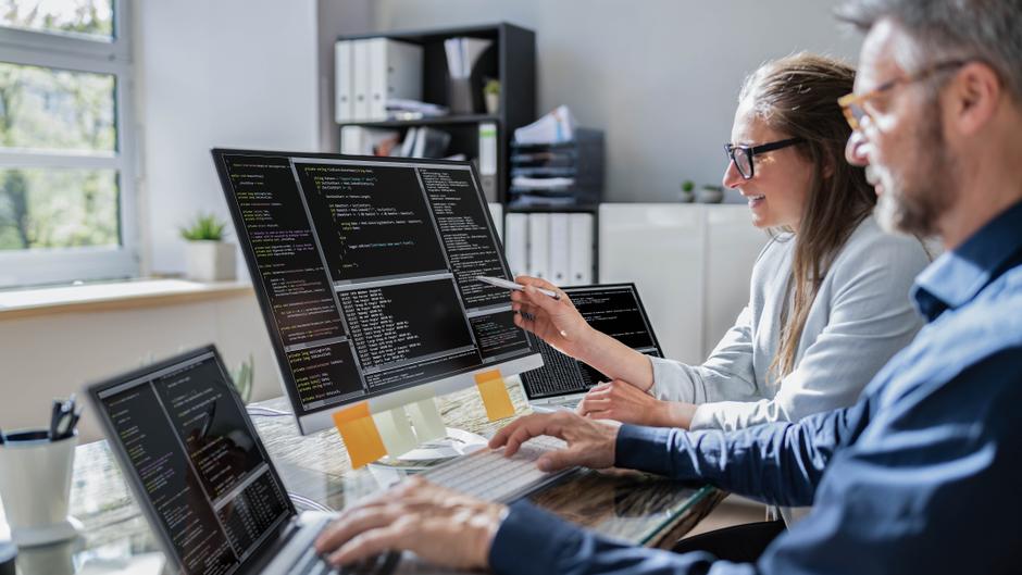Zwei Personen arbeiten an mehreren Computern, die Programmiercode auf Bildschirmen anzeigen.