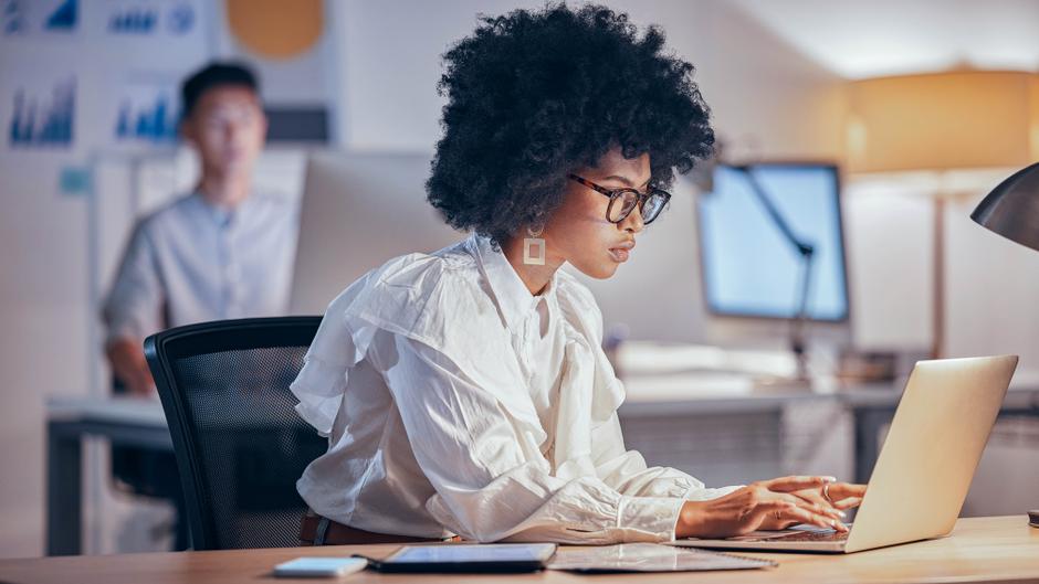 Eine Person mit lockigem Haar und Brille arbeitet konzentriert an einem Laptop in einem modernen Büro.