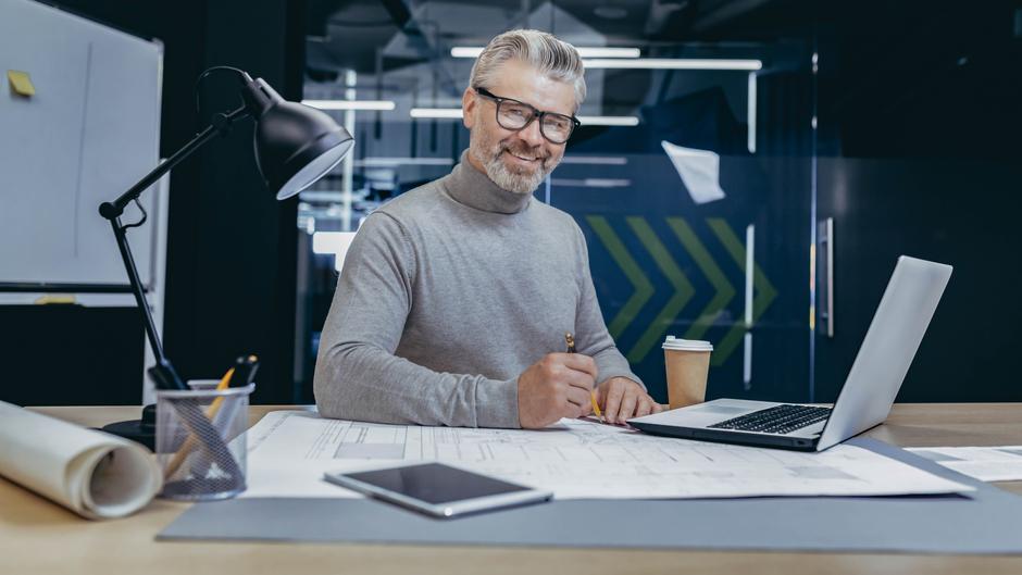 Ein älterer Mann mit Brille sitzt an einem Tisch, schreibt auf Plänen und hat einen Laptop sowie eine Kaffeetasse vor sich.