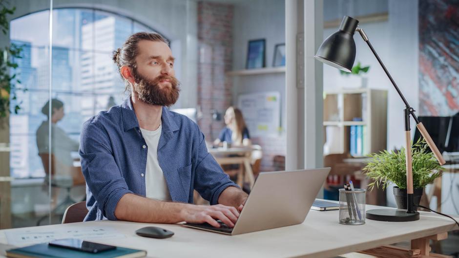 Mann mit Bart arbeitet an einem Laptop in einem modernen Büro, im Hintergrund sind weitere Personen und Pflanzen zu sehen.