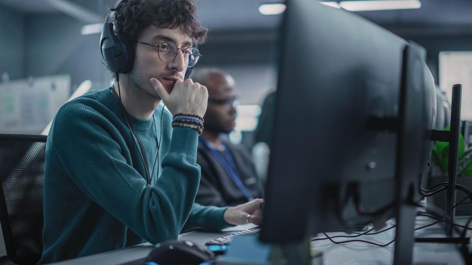 Junger Mann mit Kopfhörern sitzt vor einem Computer und schaut konzentriert auf den Bildschirm.