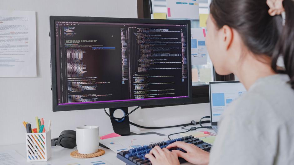 Eine Person sitzt am Schreibtisch und tippt auf einer Tastatur, während sie Code auf einem großen Monitor betrachtet.
