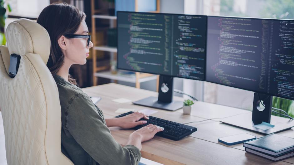 Eine Person sitzt an einem Schreibtisch und tippt auf einer Tastatur, während sie auf zwei Computerbildschirme mit Code schaut.