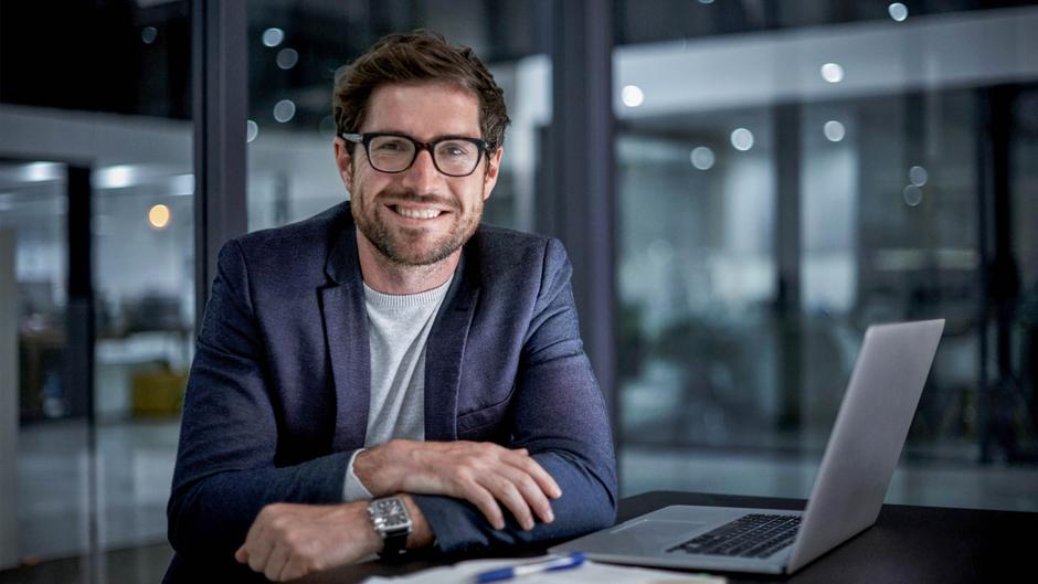 Mann mit Brille und Anzug sitzt an einem Tisch mit Laptop und lächelt in einem modernen Büro.