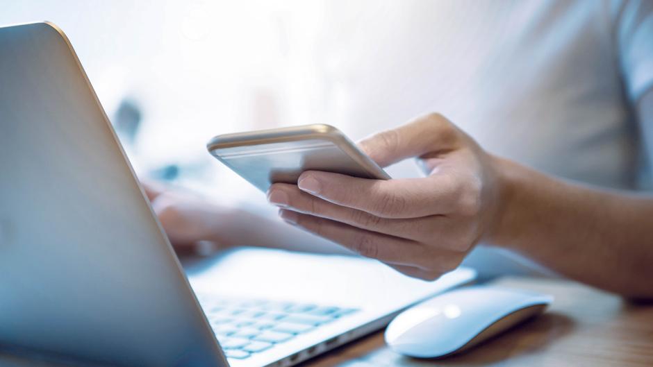 Eine Hand hält ein Smartphone, während eine andere Hand auf einem Laptop tippt.