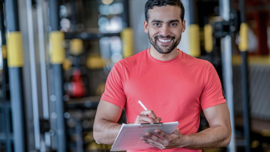 Mann in rotem T-Shirt hält ein Notizblatt und schreibt in einem Fitnessstudio.