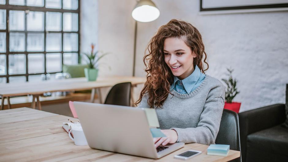 Junge Frau mit lockigem Haar sitzt an einem Tisch und arbeitet an einem Laptop in einem modernen Café.