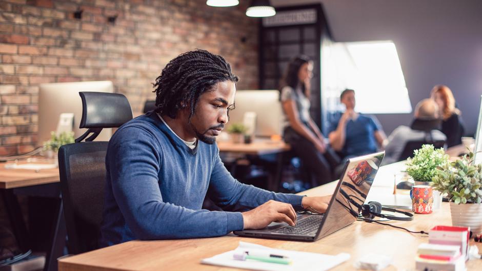 Mann mit lockigem Haar arbeitet konzentriert an einem Laptop in einem modernen Büro mit Pflanzen und Kollegen im Hintergrund.