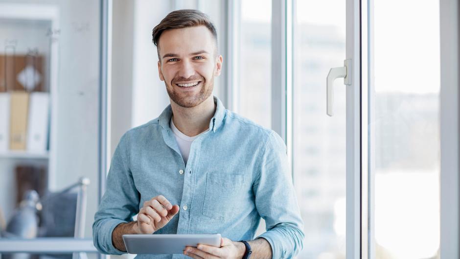 Lächelnder Mann hält ein Tablet und steht vor einem Fenster in einem modernen Büro.