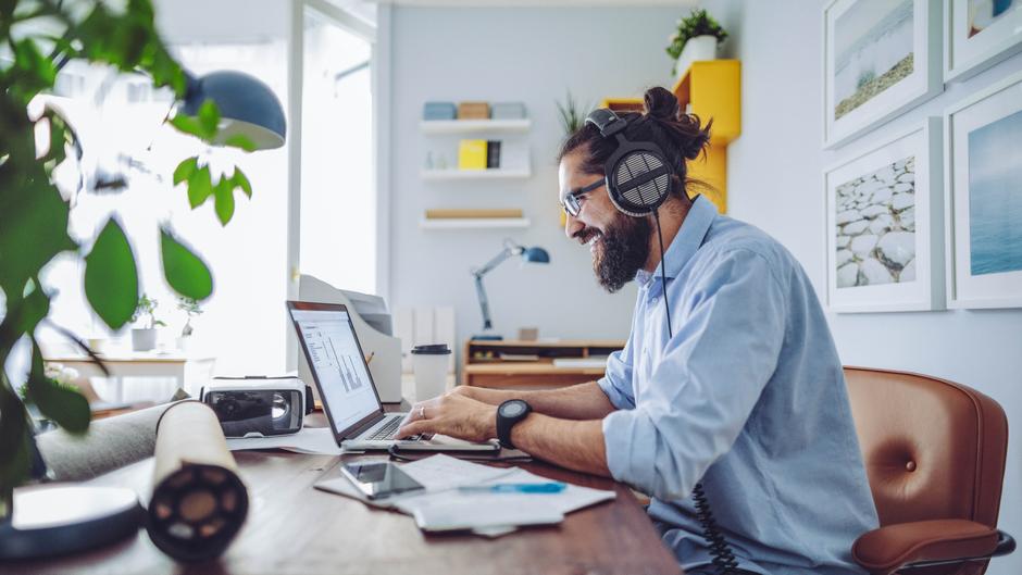 Mann mit Kopfhörern arbeitet an einem Laptop an einem Schreibtisch mit Pflanzen und Büromaterialien.