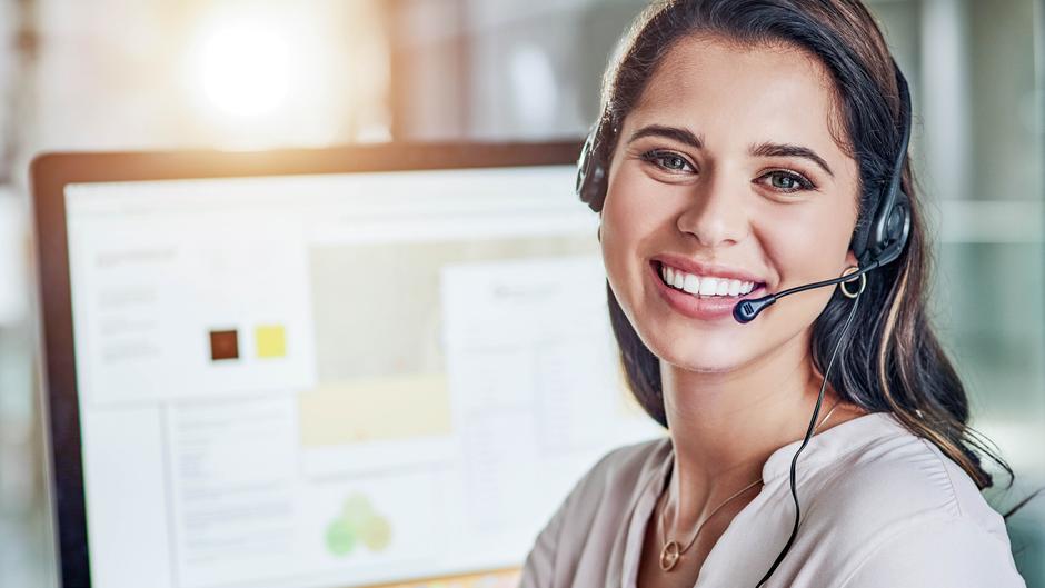 Frau mit Headset lächelt vor einem Computerbildschirm mit Grafiken und Text.